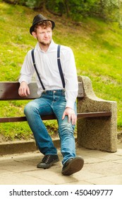 Fashion. Young fashionable man retro style wearing denim pants with straps sitting on bench in park outdoor