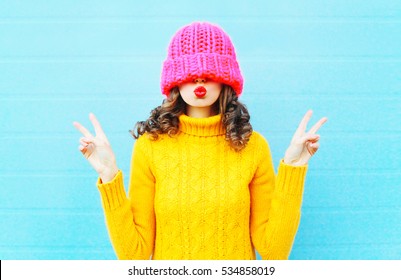 Fashion young woman blowing red lips makes air kiss wearing knitted pink hat yellow sweater over blue background - Powered by Shutterstock