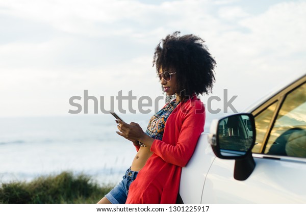 Fashionable afro hair woman on vacation texting on\
smartphone towards the sea. Stylish black model relaxing on a car\
trip to the coast.