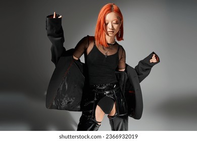 fashionable asian woman with red hair posing in blazer and black latex on grey background, chic