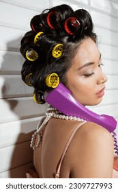 fashionable and asian young woman with hair curlers and pearl necklace talking on purple retro phone near white tiles, housewife, retro fashion, vintage-inspired