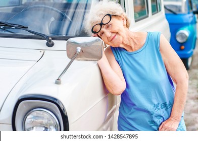 Fashionable Attractive Woman In Glasses Over 60 Is Looking At Herself In The Mirror Of A Retro Car. Summer Vacation And Travel Of Older People