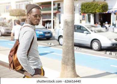 Fashionable Attractive Young Afro-American Traveler In Sunglasses And Hipster Hat Standing At Bus Stop, Waiting For Public Transport To Get To Urban Beach. Travel, Adventure, Wonderlust And Tourism