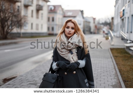 Fashionable attractive young blond woman in a vintage knitted sweater in a stylish coat in warm gloves with a vintage scarf with a fashionable black leather bag walking on the street. City cute girl.