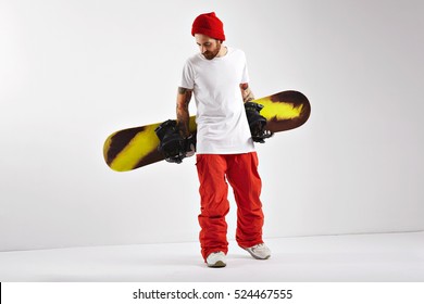 Fashionable bearded snowboarder in bright clothing and plain white cotton t-shirt holding his snowboard behind his back isolated on white