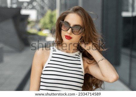 Fashionable beautiful Caucasian young woman model with red lips in stylish sunglasses in a fashion summer striped top walks in the city and straightens her hair