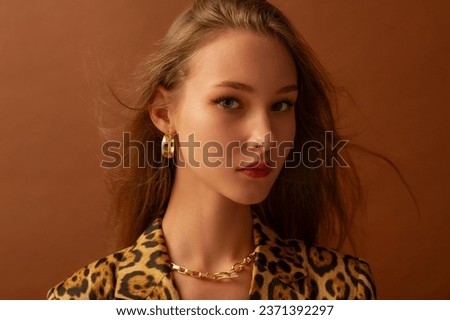 Fashionable beautiful confident woman wearing trendy golden hoop earrings, chain necklace, leopard print blazer, posing in studio, on brown background. Copy, empty space for text