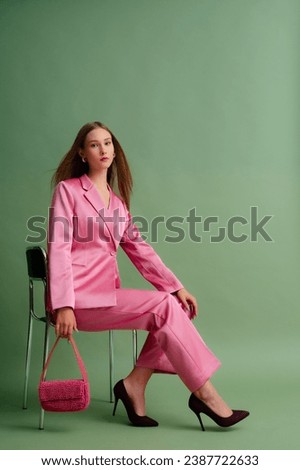 Fashionable beautiful confident woman wearing trendy pink satin suit blazer, trousers, pointed toe high-heeled shoes, holding beaded bag, posing on green background. Full-length studio portrait