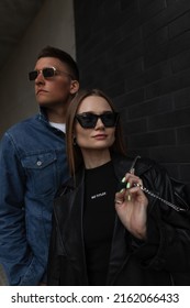 Fashionable Beautiful Couple In Stylish Street Fashion Outfit With Jacket, Bag And Sunglasses Walking On The Street On A Dark Background. Woman's And Man's Urban Casual Outwear