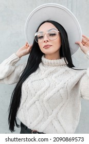 Fashionable beautiful girl with stylish glasses in a vintage knit  sweater wears a hat near a gray wall on the street and looks at the camera