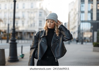 Fashionable beautiful teenager woman in stylish clothes with a hat, black sweater and leather jacket walks in the city