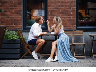 Fashionable Beautiful Young Couple Sitting In Cafe And Drinking Coffee Outside Near The House. Natural Color. Stylish Woman Feeding With Spoon Man In City Coffee Shop. Lifestyle Full Length Portrait