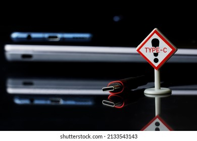 Fashionable black and red usb type-c plug next to thin laptop, smartphone and type-c sign. Black background. A modern way to transfer data and charge gadgets. Selective focus. Macro.