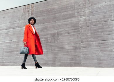 Fashionable black woman in red coat walking on out of focus background. Stylish African American woman in bright red jacket, black pants and handbag walking down the street. copy space photography