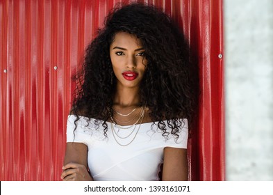 Fashionable black woman with red lips leaning in a red wall posing on the street while looking at camera
