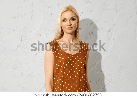 Fashionable blonde woman standing indoors in brown sleeveless blouse with dotted pattern
