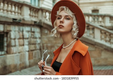 Fashionable blonde woman wearing orange hat, trench coat, pearl necklace, ring, posing in street of European city. Copy, empty space for text