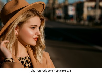 Fashionable blonde woman wearing trendy beige hat, coat, long earrings, posing in street of European city. Copy, empty space for text