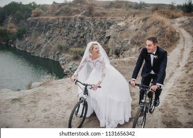 A Fashionable Bridegroom In A Black Suit And A Beautiful Bride In A Lace Dress With A Long Veil Ride On Bicycles And Laugh At The Background Of Rocks And Stones. Autumn Wedding Portrait Of Gay.