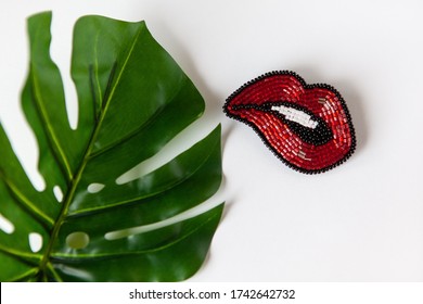 Fashionable brooch in shape of lips from Japanese beads, green leaf of Monstera on white background. Close-up, flat lay, copy space. Concept trends, fashion, style, DIY, needlework