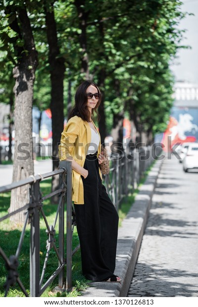 Fashionable brunette woman in a yellow jacket and\
black pants, sunglasses and bag string bag walk on the street.\
Fashion spring summer\
photo.