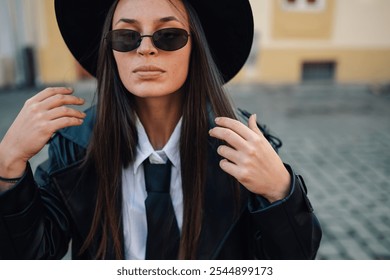 Fashionable businesswoman adjusting her long brown hair, wearing a black hat, sunglasses, white shirt, and tie, creating a chic and androgynous look - Powered by Shutterstock