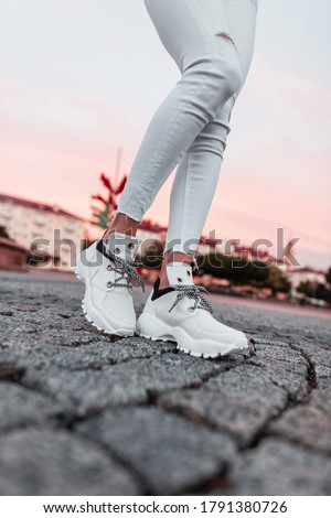 Fashionable casual white sneakers on female legs on a stone road at pink sunset. Close-up.