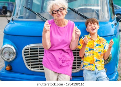 Fashionable, Cheerful Grandmother, Grandchild Tourists Are Fooling Around A Retro Car Imitating Driving. Summer Family Traveling. Laughing Elderly Woman And A Child Are Playing Drivers. Travel Concept