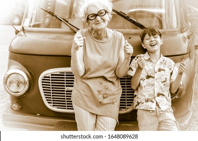 Fashionable, Cheerful Grandmother With Her Grandchild Are Fooling Around A Retro Car Imitating Driving. Summer Family Vacation And Travel. Retro Style. Sepia