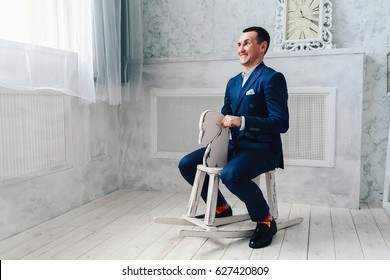 Fashionable Cheerful Man In A Blue Suit Is Sitting On A Toy Horse. Adult Acting Like A Child Concept.