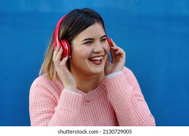 Fashionable Chubby Woman In Pink Cardigan Listening To Music With Red Headphones And Smiling Looking Away From The Camera - Body Positivity And Self Esteem Concept