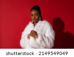 Fashionable confident Black woman wearing luxury white faux fur coat, trendy long earrings with rhinestones,  posing on red background. Copy, empty space for text