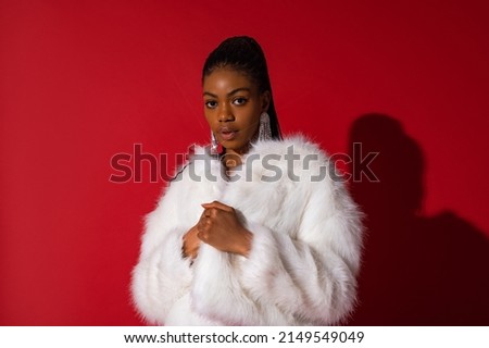 Fashionable confident Black woman wearing luxury white faux fur coat, trendy long earrings with rhinestones,  posing on red background. Copy, empty space for text