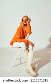 Fashionable confident blonde woman wearing trendy orange sweatshirt, color sunglasses, white skinny jeans, high leather boots, posing on white background. Copy, empty space for text - Shutterstock ID 2117571815