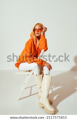 Fashionable confident blonde woman wearing trendy orange sweatshirt, color sunglasses, white skinny jeans, high leather boots, posing on white background. Copy, empty space for text