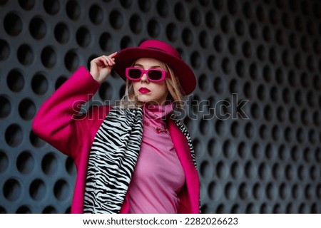 Fashionable confident blonde woman wearing trendy fuchsia color hat, coat, rectangular sunglasses, pink turtleneck, zebra print scarf, posing in street of city. Copy, empty space for text
