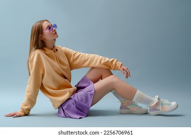 Fashionable confident girl wearing purple sunglasses, pleated mini skirt, yellow hoodie, socks, high top colorful sneakers, sitting, posing on blue background. Full-length studio portrait. Copy space