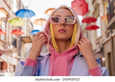 Fashionable confident girl wearing trendy pink sunglasses with yellow plastic chain, hood, blue trench coat, posing in European city. Streetstyle, street fashion conception. Close up portrait