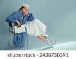 Fashionable confident woman wearing beret, elegant blue coat, white denim maxi skirt, silver ankle boots, holding stylish faux leather purse, posing on blue background. Full-length studio portrait