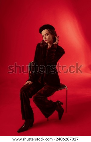 Fashionable confident woman wearing beret, velvet suit blazer, wide trousers, leather ankle boots, sitting on chair, posing on red background. Full-length studio fashion portrait