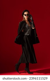  Fashionable confident woman wearing black cat eye sunglasses, leather beret, gloves, waistcoat, boucle blazer, pleated midi skirt, high heel sock boots, posing on red background. Full-length portrait