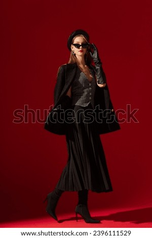  Fashionable confident woman wearing black cat eye sunglasses, leather beret, gloves, waistcoat, boucle blazer, pleated midi skirt, high heel sock boots, posing on red background. Full-length portrait
