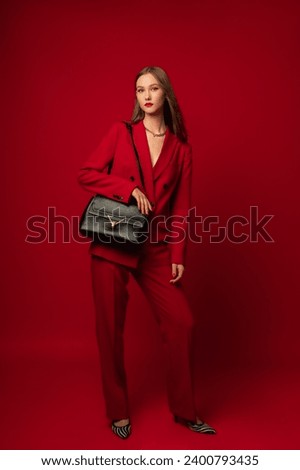 Fashionable confident woman wearing classic red suit double breasted blazer, trousers, zebra print shoes, with black faux leather bag, posing on red background. Full-length studio fashion portrait