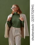 Fashionable confident woman wearing elegant white suit with blazer, trousers, cashmere turtleneck sweater, trendy cat eye sunglasses, posing on green background. Studio fashion portrait