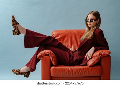 우아한 마르살라 색 정장을 입고, 선글라스를 끼고, 표범의 프린트 로퍼 신발을 신고, 포즈를 취하고, 갈색 가죽 안락의자에 앉아 있는 세련된 자신감 넘치는 여성. 스튜디오 패션 초상화 복사 텍스트의 빈 공간 스톡 사진