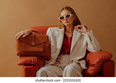 Fashionable confident woman wearing elegant white suit, sunglasses, chunky chain, holding classic brown leather bag, sitting in armchair, posing on beige background. Copy, empty space for text - Shutterstock ID 2255335105