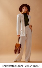 Fashionable confident woman wearing elegant white suit with blazer, wide leg trousers, turtleneck top, hat, leather ankle boots, holding brown suede fringed bag,  posing on beige background - Shutterstock ID 2255441267