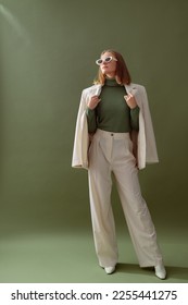 Fashionable confident woman wearing elegant white suit with blazer, wide leg trousers, cashmere turtleneck sweater, trendy sunglasses, posing on green background. Full-length studio fashion portrait - Shutterstock ID 2255441275