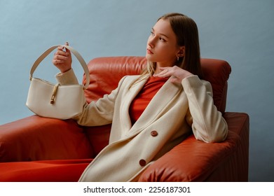 Fashionable confident woman wearing elegant white woolen coat,  holding stylish leather bag, sitting in armchair, posing on blue background. Copy, empty space for text