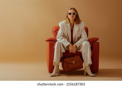 Fashionable confident woman wearing elegant white suit, sunglasses, ankle boots, holding classic brown leather bag, sitting in armchair, posing on beige background. Copy, empty space for text - Shutterstock ID 2256187303